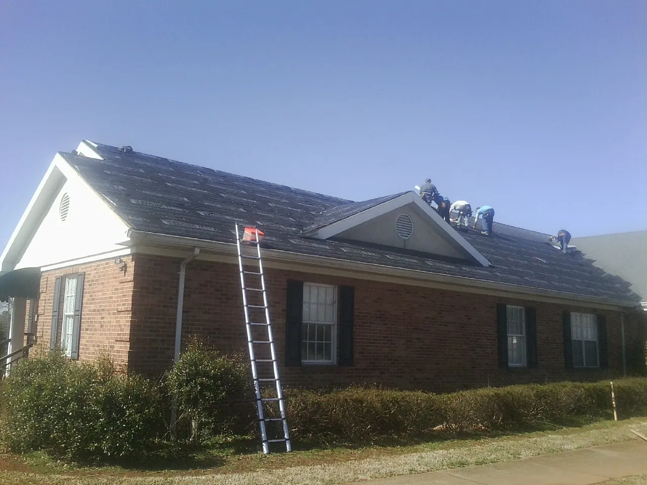 Crew tearing shingles off a brick ranch roof with a ladder set — Emergency Roof Repair work by our crew in Cusseta