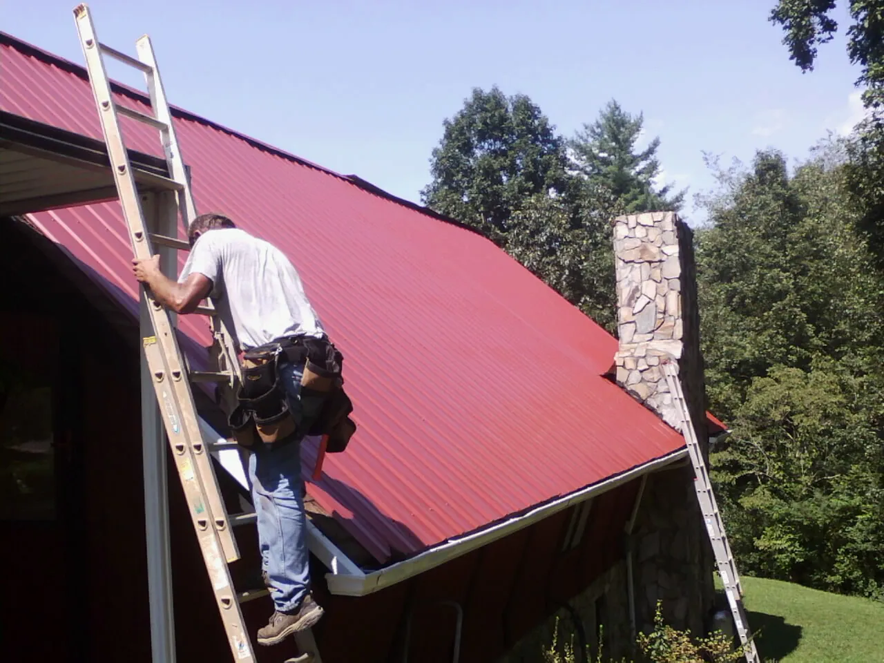 Roofer on a ladder beside a red metal roof and stone chimney — Soffit & Fascia Repair by Roofing Brothers in Cusseta